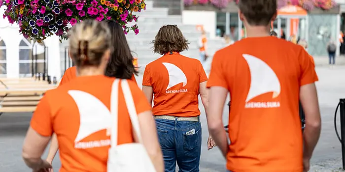 Ryggen til tre mennesker som har oransje t-skjorter med logoen til Arendalsuka på. Foto: Mona Hauglid