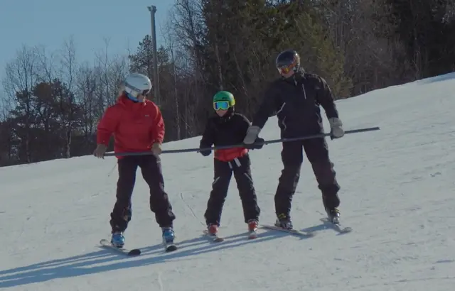 Ett barn og to voksne side om side ned slalåmbakken - med en statur mellom seg