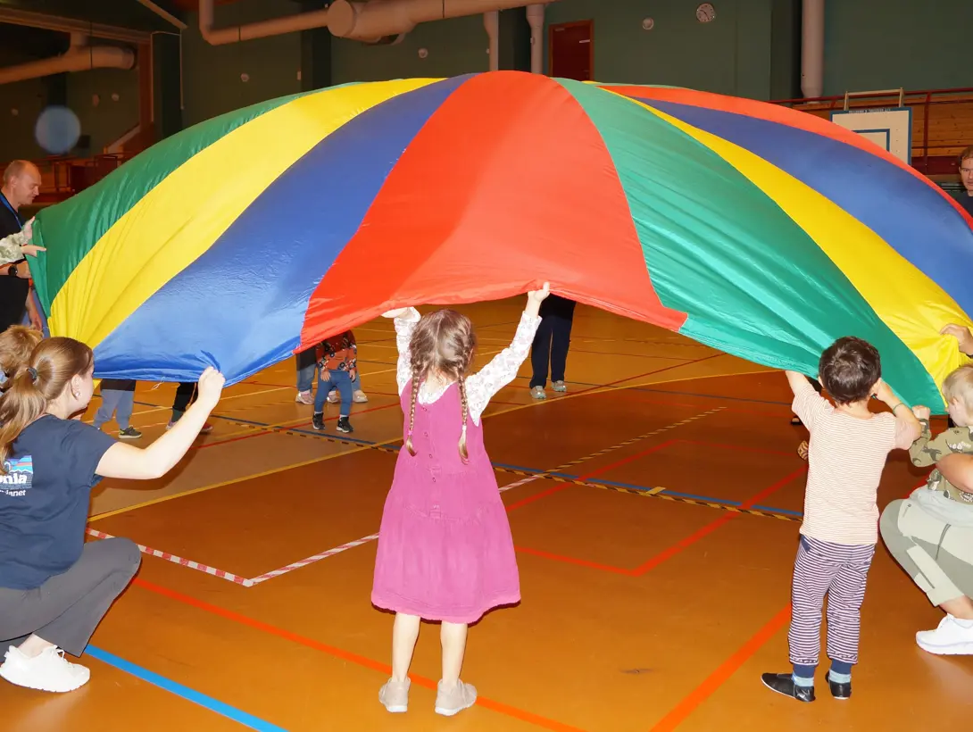 En gruppe barn og voksne leker med den fargerike fallskjermen